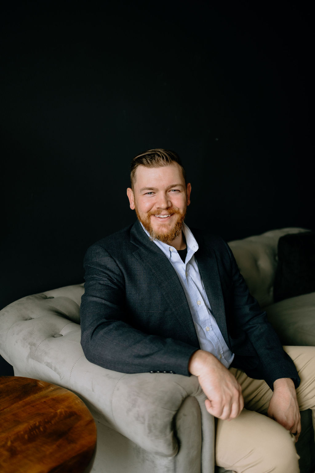 Joshua Schafer seated on a gray tufted sofa, wearing a navy blazer, smiling warmly at the camera