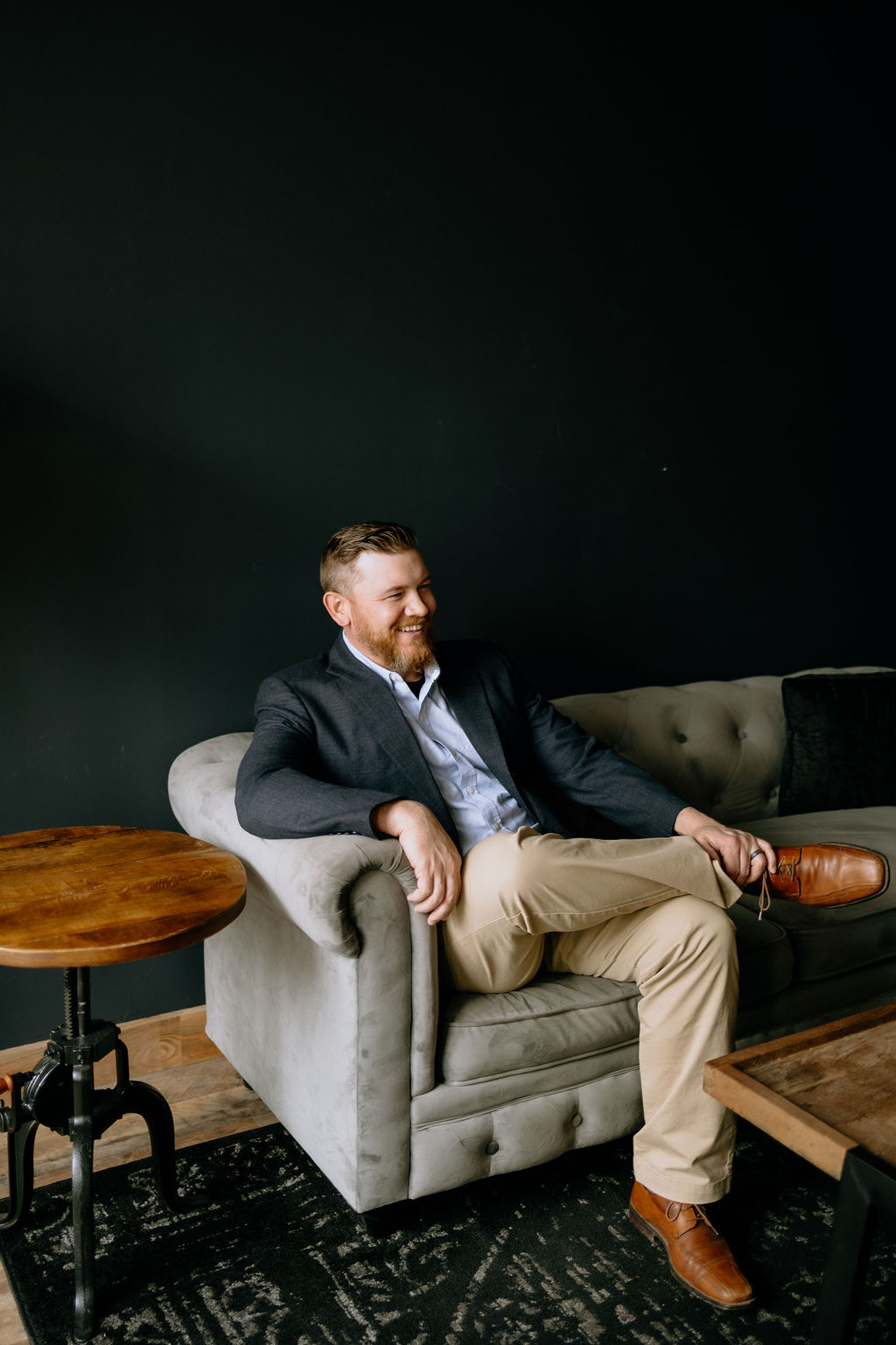 Joshua Schafer laughing and relaxed, seated on a gray sofa in a dark studio setting