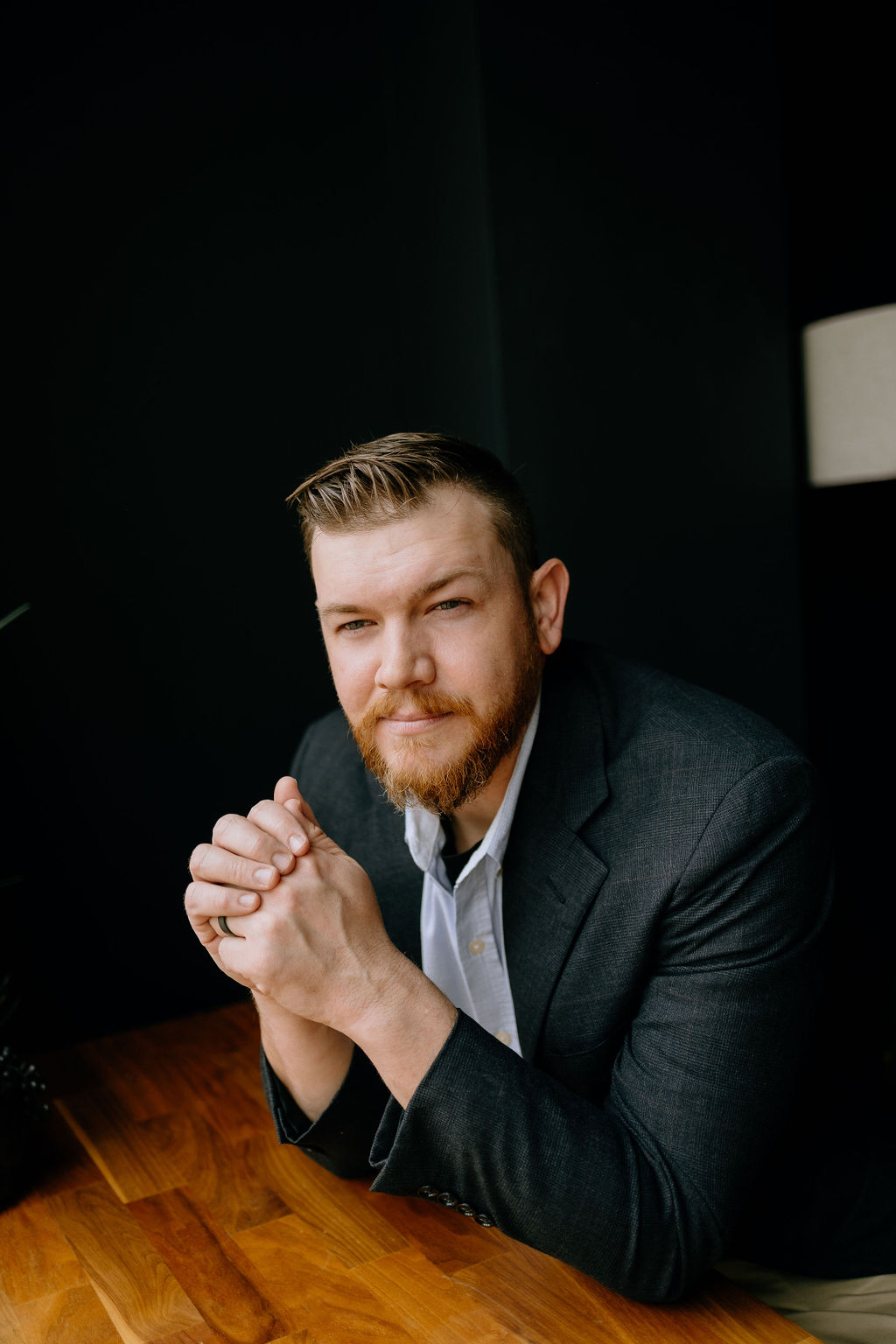 Joshua Schafer leaning forward at a wooden table with hands clasped together, looking directly into the camera with a focused, thoughtful expression