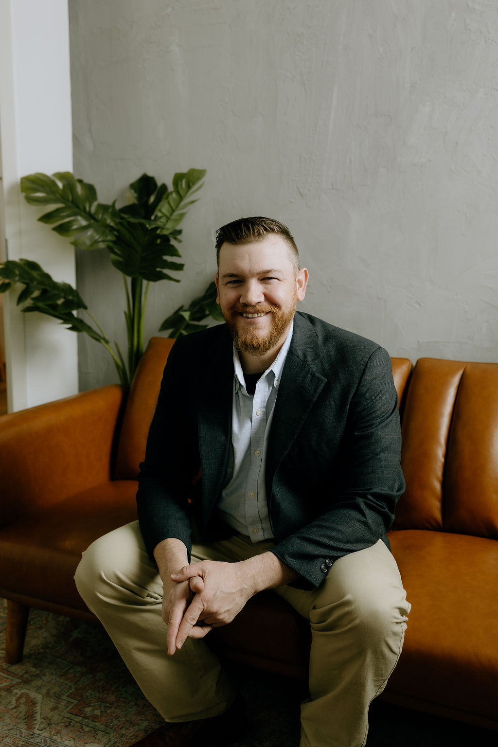 Joshua Schafer seated on a tan leather sofa with a large green plant in the background, smiling confidently