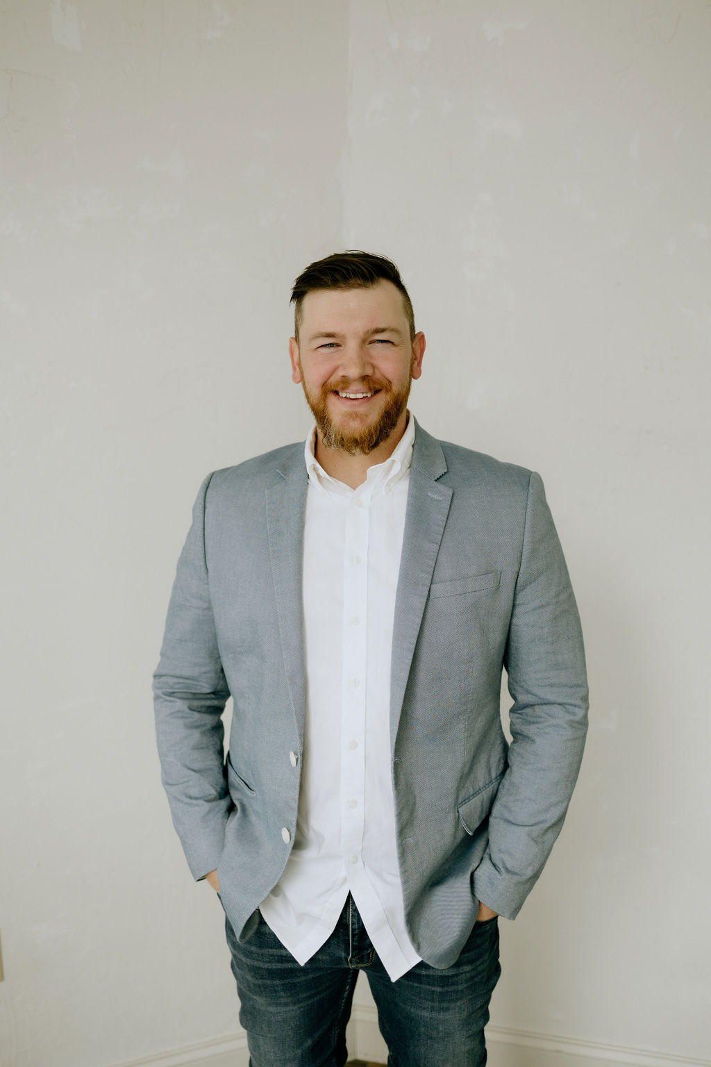 Joshua Schafer standing in a light gray blazer and jeans, smiling with hands relaxed at his sides
