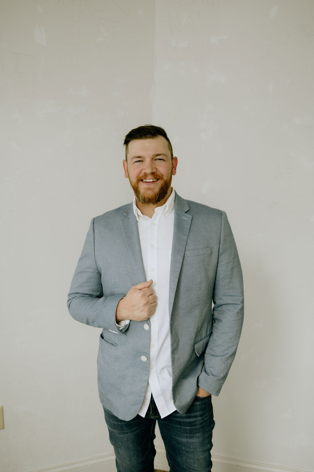 Joshua Schafer standing in a light gray blazer, smiling with one hand resting on his lapel