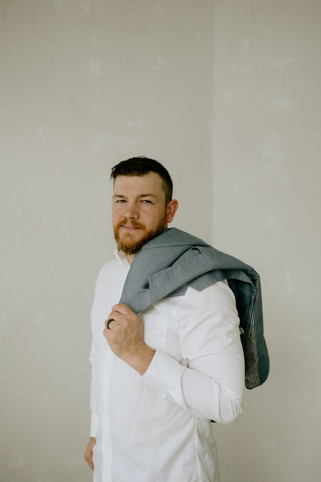 Joshua Schafer in a white button-down shirt, holding a gray blazer over his shoulder, looking back with a calm confident expression